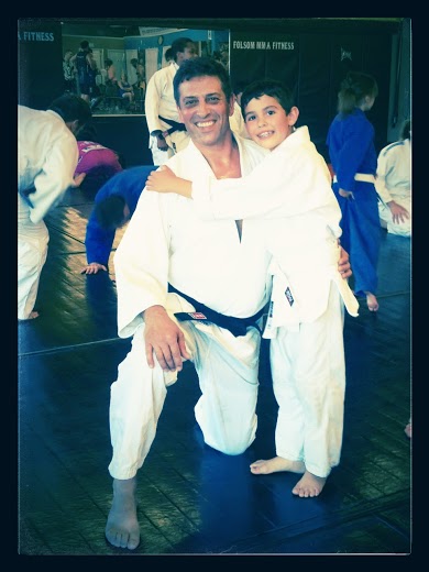 A coach kneeling on the mat with his arm around a young student, both smiling